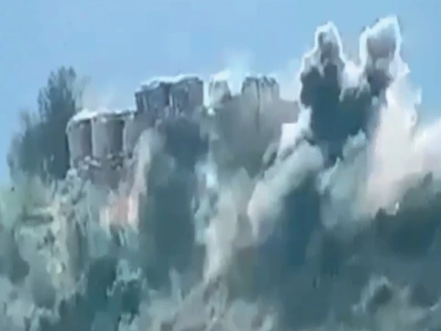 Smoke and dust rising from a hilltop border post after artillery fire, seen across a wooded valley