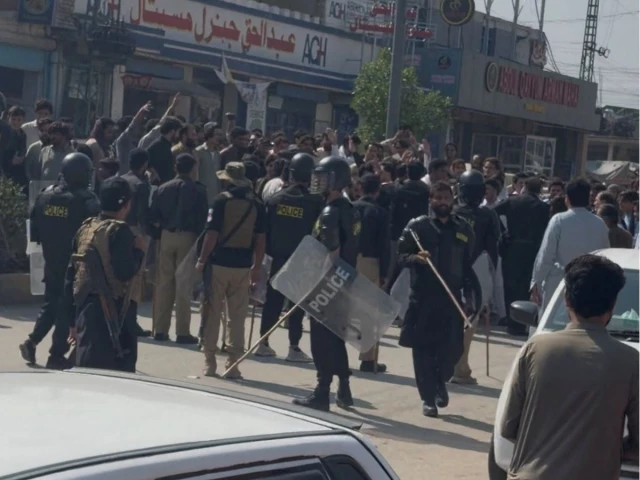 Riot police with shields and batons facing a crowd of protesters on a commercial street in Bara